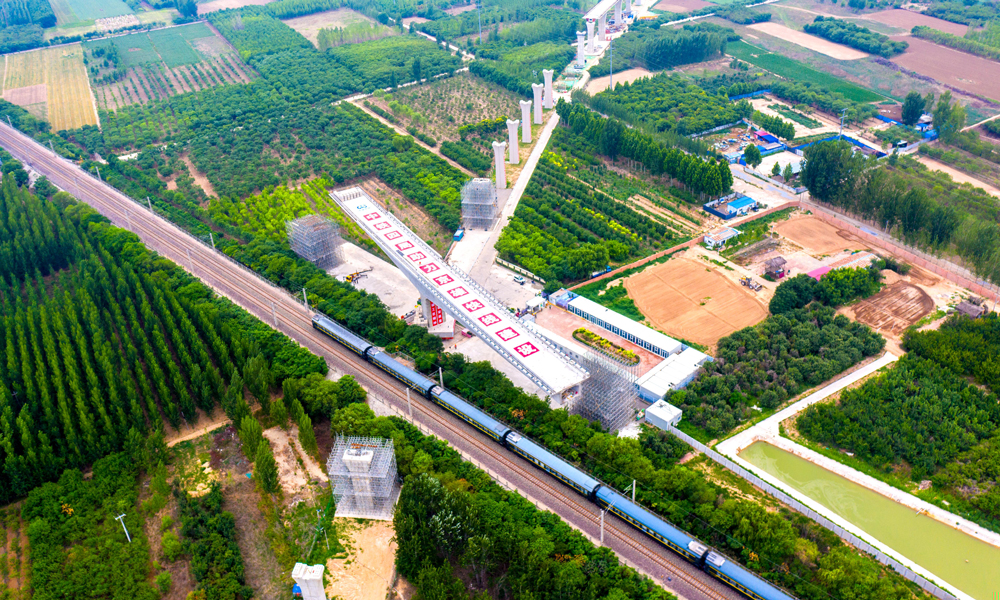 跨京九鐵路連續(xù)梁是雄安機場快線首座涉及運營鐵路的轉體梁，所跨京九鐵路每日運行列車多達百余列。為確保轉體期間京九鐵路安全運營，中鐵四局多次組織專家評審、優(yōu)化施工方案，并在正式轉體前試轉，通過稱重平衡試驗測試轉體部分的不平衡力矩、偏心矩、摩阻力矩及摩擦系數等參數，以實現橋梁轉體的配重要求，在轉體過程中，根據轉臺上標識的轉角刻度及梁面軸線實行嚴格監(jiān)控。圖為跨京九鐵路轉體梁轉體前航拍。