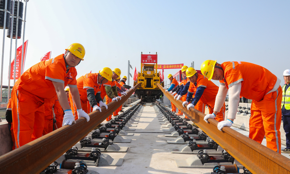 為優質高效完成鋪軌任務，建設者提前在位于荊州站北側的鋪軌基地儲存長鋼軌，以鋪軌基地為起點，采用TC-500型鋪軌機由荊州站一路向北鋪設長鋼軌；加持智能行車調度指揮系統，全天候對鋪軌機、長軌運輸車、焊軌車等設備的運行狀態及作業位置進行監控和統一調度指揮，保證了交叉施工安全有序，并且提高了鋪軌速度，每天單向鋪軌可達6公里。圖為鋪軌作業現場，作業人員在鋪軌機的牽引下正在安裝軌道。